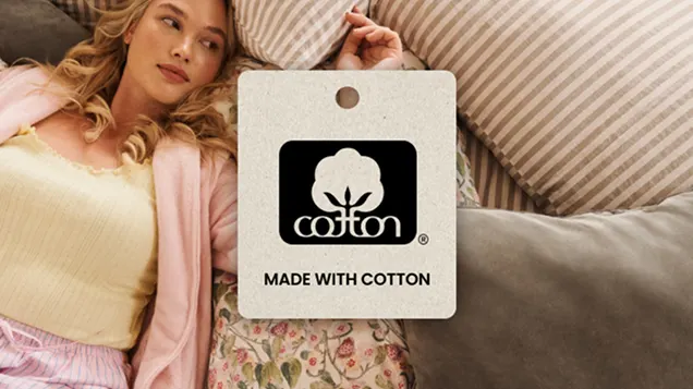 Close-up of a 'Made with Cotton' tag placed in front of a woman lounging on a couch with cotton pillows and floral cotton clothing.
