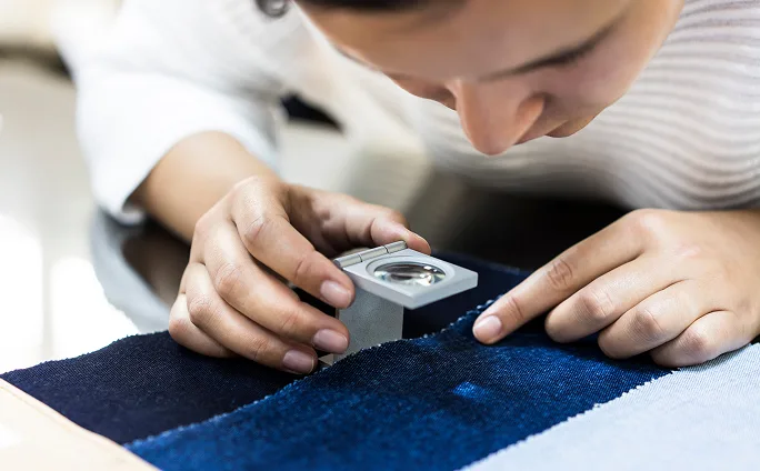 Close-up of a person inspecting a piece of cotton fabric with a magnifying glass.