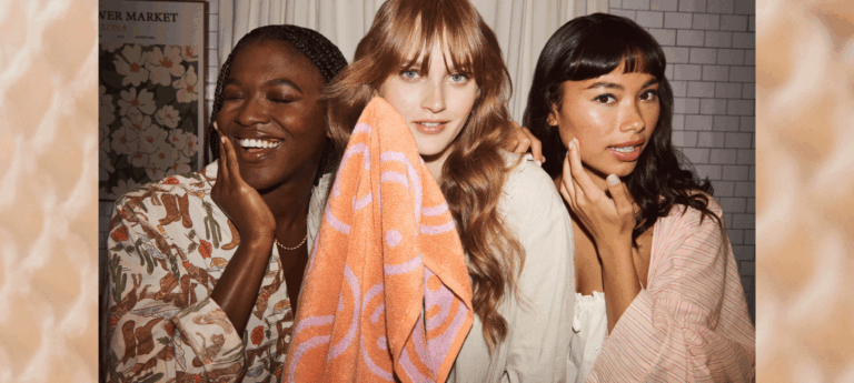 three women standing in front of mirror using cotton products to remove makeup